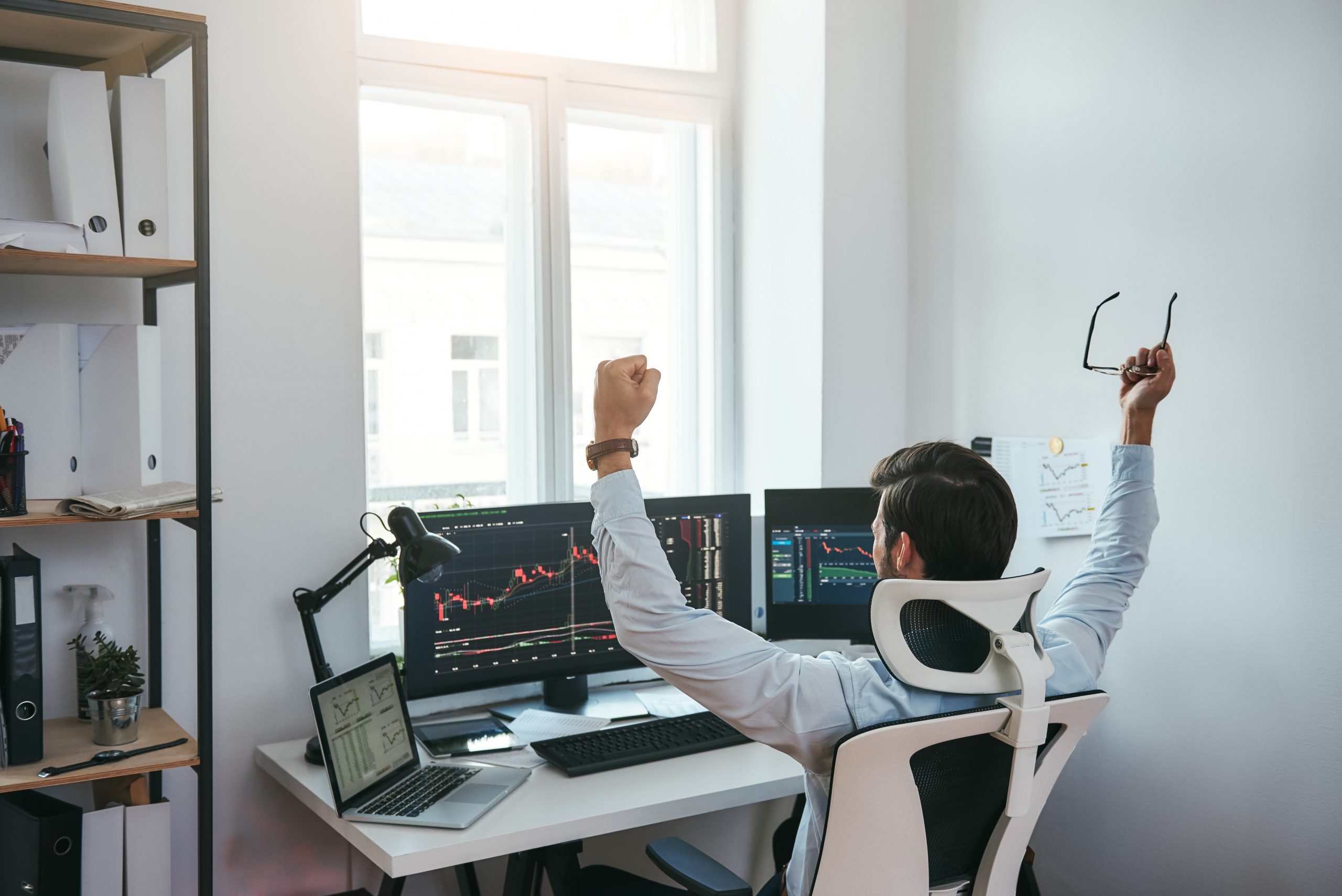Winner Back view of stock trader with raised hands holding eyeglasses and feeling happy while looking at multiple computer screens with data and charts in his modern office.